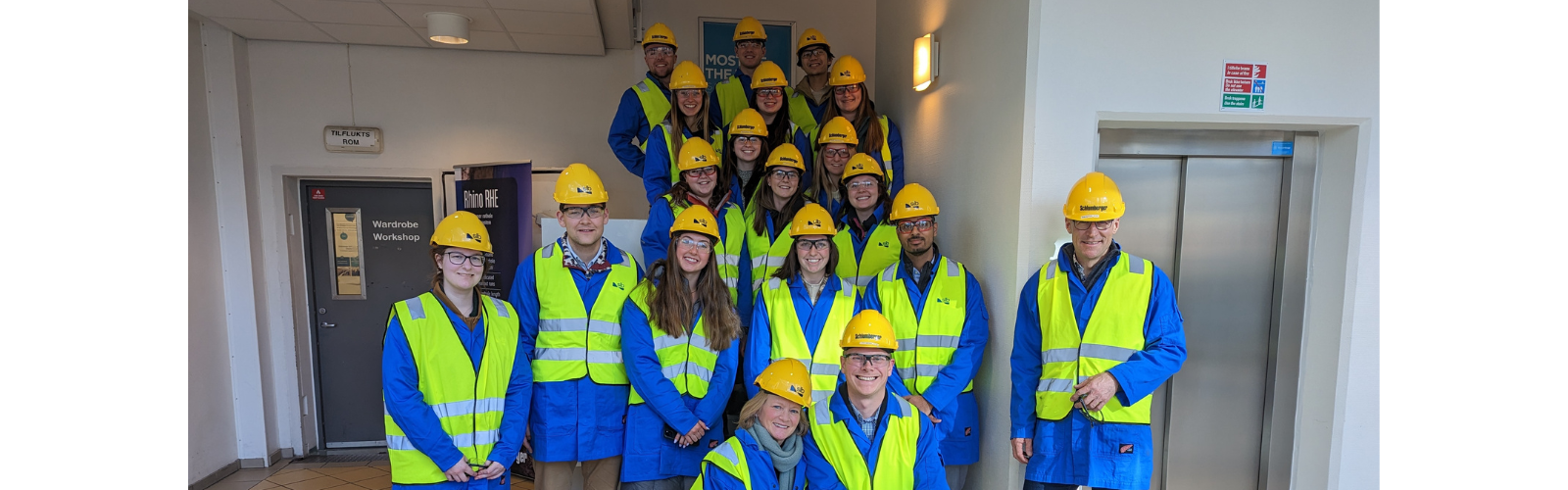 Several students stand smiling in a stairwell, wearing blue protective clothes, yellow high-vis vests, safety glasses and yellow hard hats. 